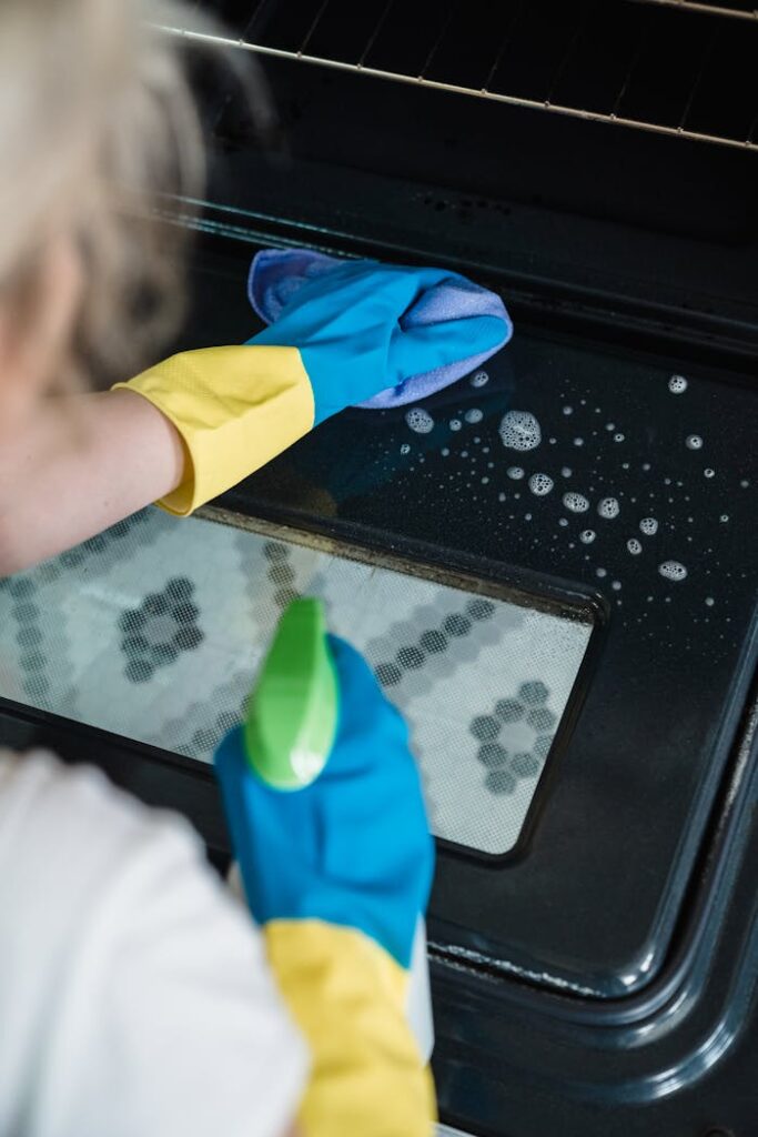 pexels-photo-9462315 A person wearing gloves cleans an oven door using a spray and cloth.